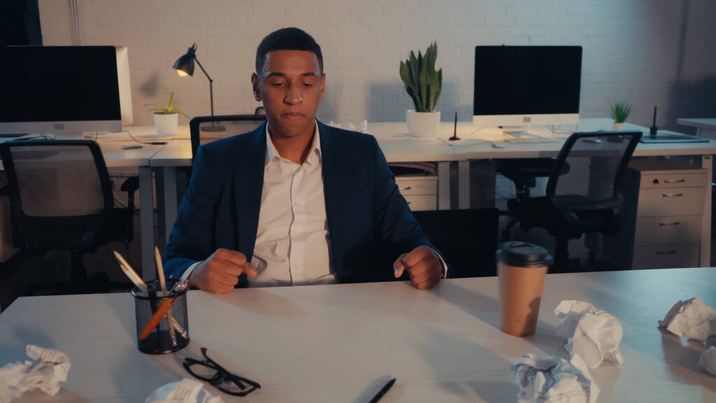 Arrabbiato uomo d'affari afroamericano in giacca e cravatta guardando carta accartocciata vicino al caffè per andare in ufficio la sera  - Foto, immagini