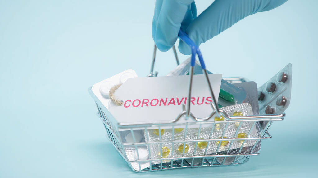 vista parziale della persona in guanto di lattice che tiene il carrello della spesa con coronavirus lettering su carta e medicamenti su blu   - Foto, immagini