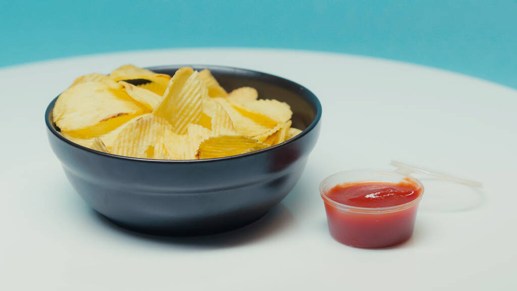 batatas fritas crocantes e enrugadas em tigela perto de recipiente de plástico com ketchup em azul - Foto, Imagem