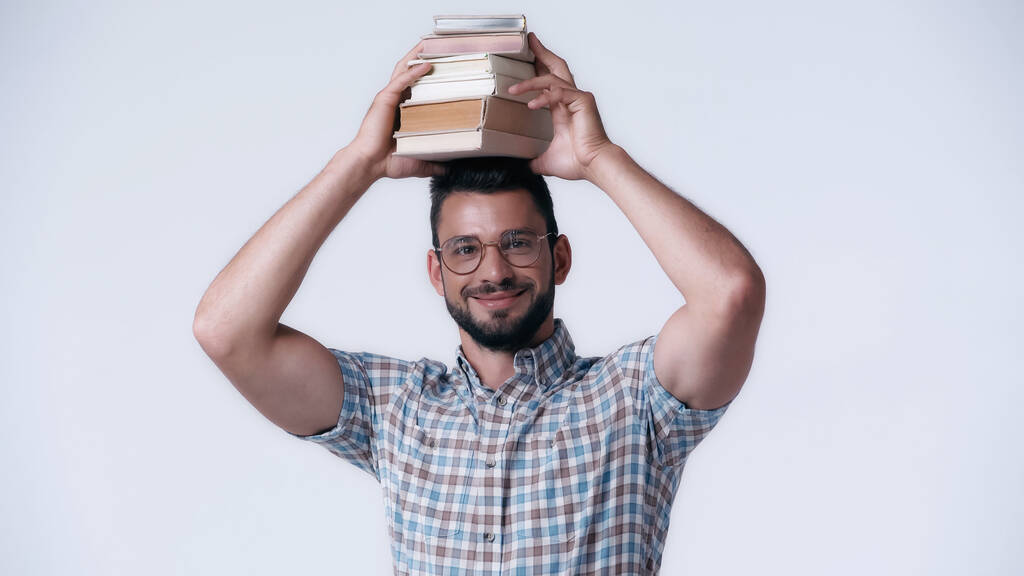 веселый ботаник в очках держит стопку книг над головой изолированных на сером - Фото, изображение