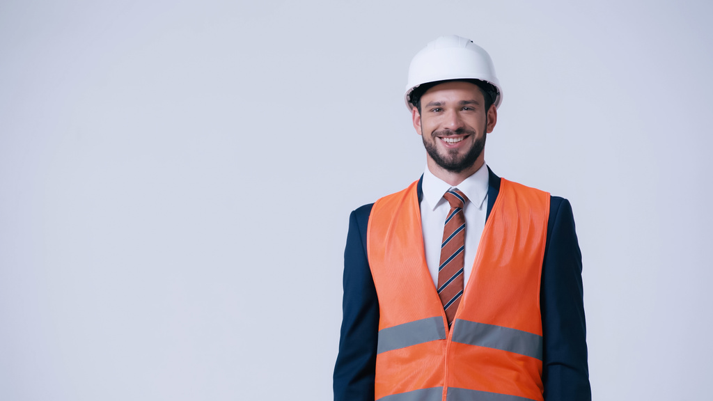 smiling engineer in hardhat and safety vest looking at camera isolated on grey - Photo, Image