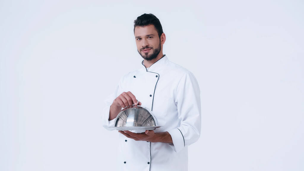 брюнетка чоловік в куртці шеф-кухаря тримає тарілку з шафою і дивиться на камеру ізольовано на білому
 - Фото, зображення