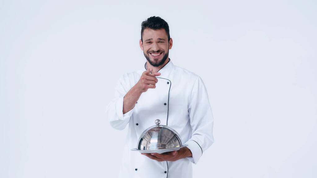 laughing chef holding serving dish with stainless cloche and pointing with finger at camera isolated on white - Photo, Image