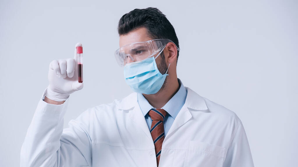 imunologista em máscara médica e óculos olhando para tubo de ensaio com amostra de sangue isolado em cinza - Foto, Imagem