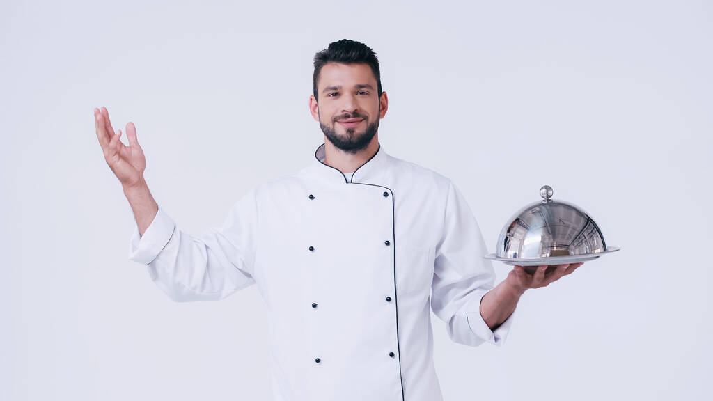 alegre chef gesto mientras sostiene servir plato con cloche aislado en blanco - Foto, imagen