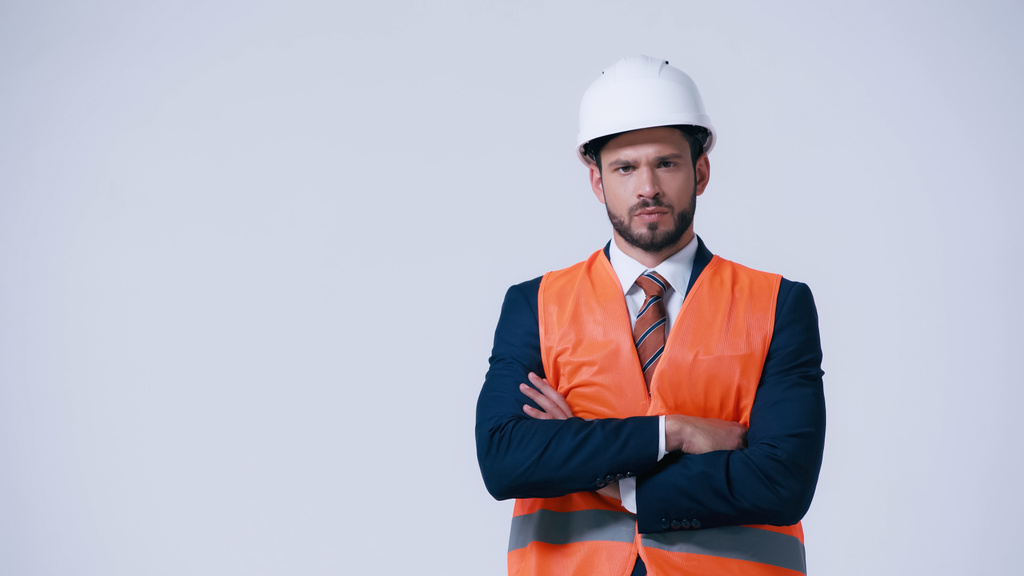 строгий инженер в защитном жилете и шлеме, стоящий со скрещенными руками и смотрящий на камеру, изолированную на сером - Фото, изображение