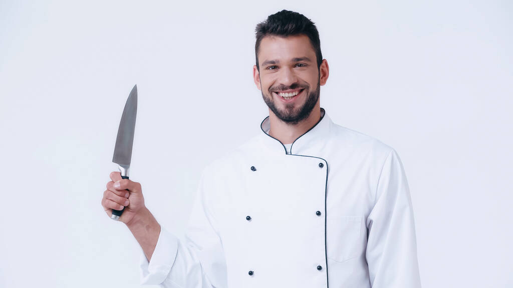 alegre chef en uniforme sosteniendo cuchillo afilado y mirando a la cámara aislada en blanco - Foto, imagen