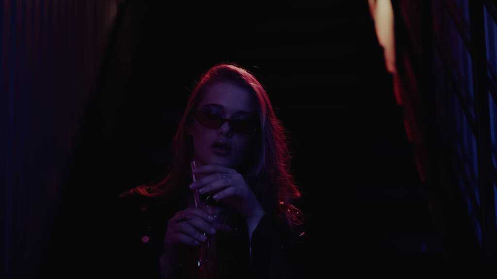Donna in occhiali da sole che tiene cocktail in nightclub scuro  - Foto, immagini