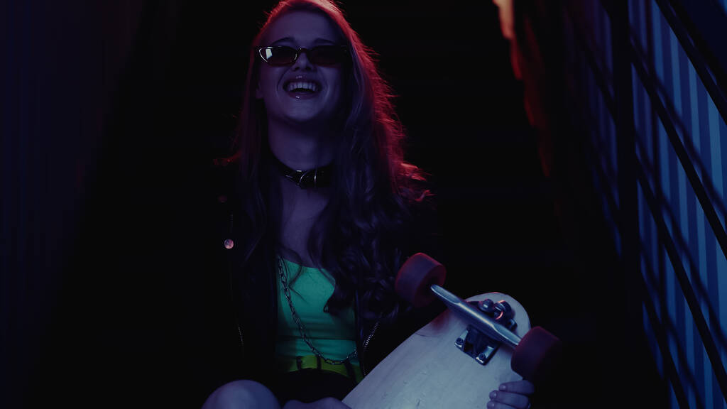 Fröhliche Frau mit Sonnenbrille hält Longboard in Nachtclub  - Foto, Bild