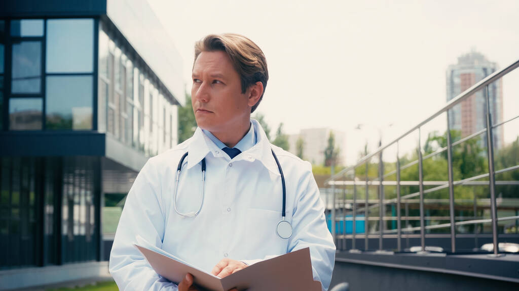 orvos fehér köpenyben sztetoszkóppal és mappával néz el a szabadban - Fotó, kép