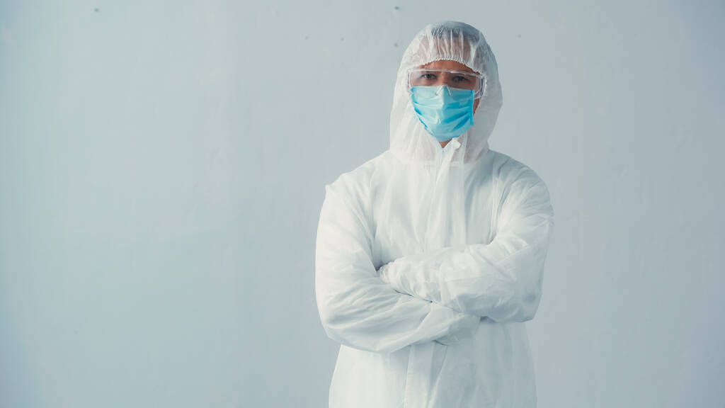 doctor in hazmat suit and medical mask with goggles standing with crossed arms on grey background - Photo, Image
