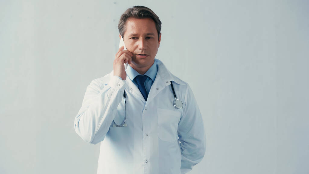 lékař v bílém plášti stojí s rukou za zády, zatímco mluví na smartphone na šedé - Fotografie, Obrázek