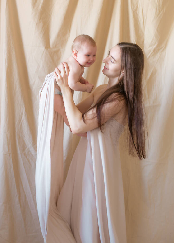Mladá matka s dlouhými tmavými vlasy drží roztomilou dcerku v náručí v nahé látce na nahém textilním pozadí. šťastné mateřství - Fotografie, Obrázek