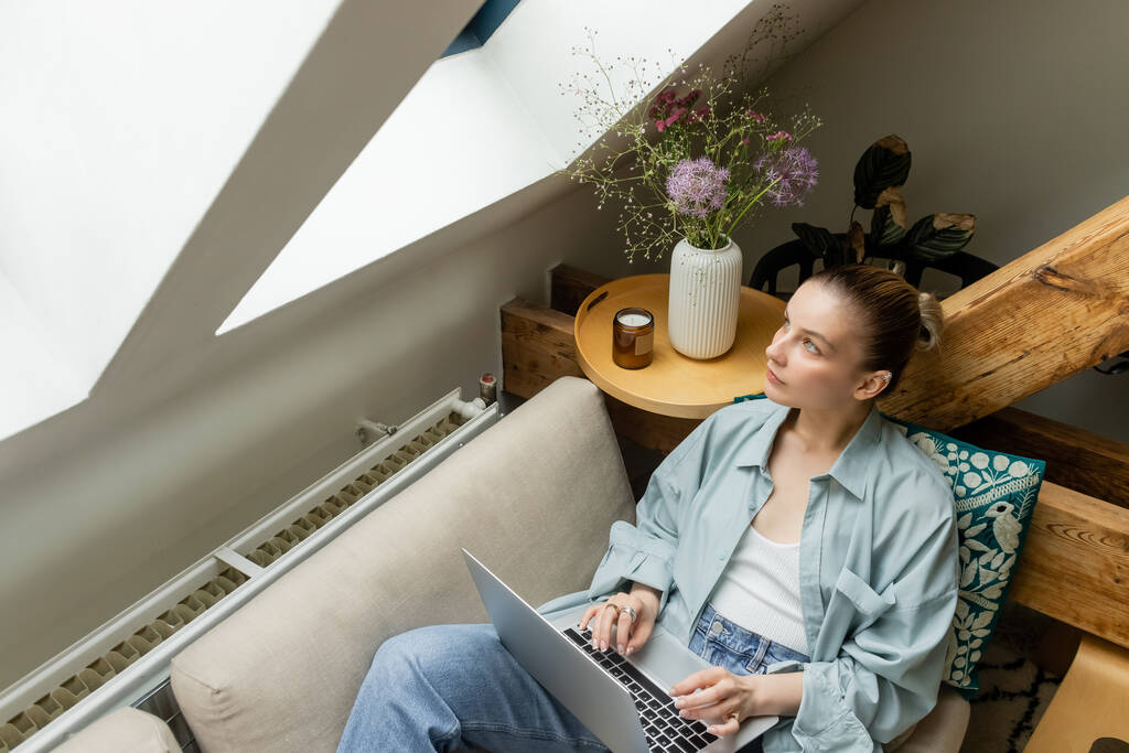 Високий кут зору жінки, дивлячись на вікно, використовуючи ноутбук на дивані вдома
  - Фото, зображення
