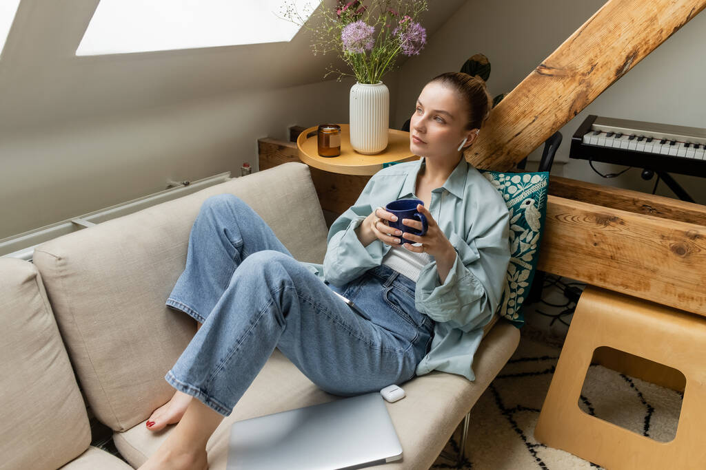 Молодая женщина в наушнике держит чашку возле ноутбука на диване дома  - Фото, изображение