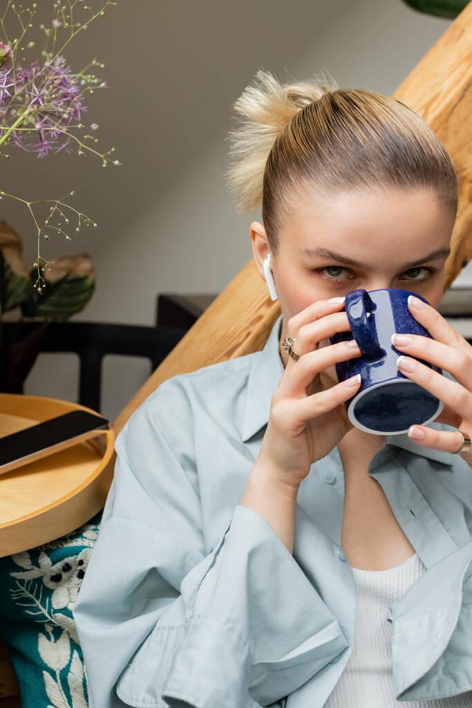 Fiatal nő fülhallgató ivás kávé közelében smartphone üres képernyő otthon  - Fotó, kép