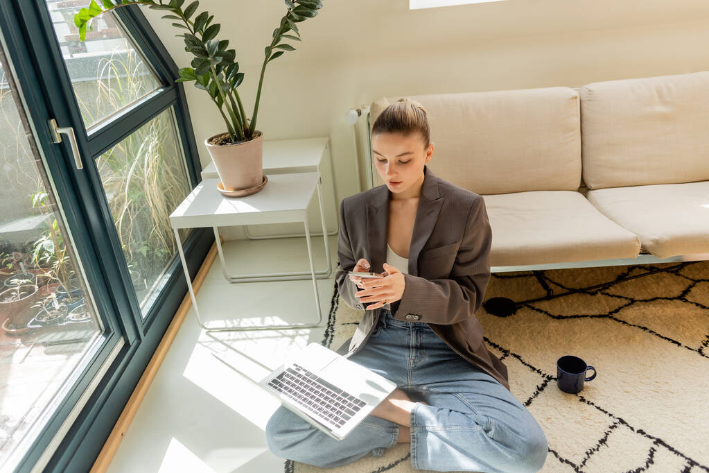 Jeune femme en veste et jeans utilisant smartphone et ordinateur portable près de la tasse sur le tapis à la maison  - Photo, image