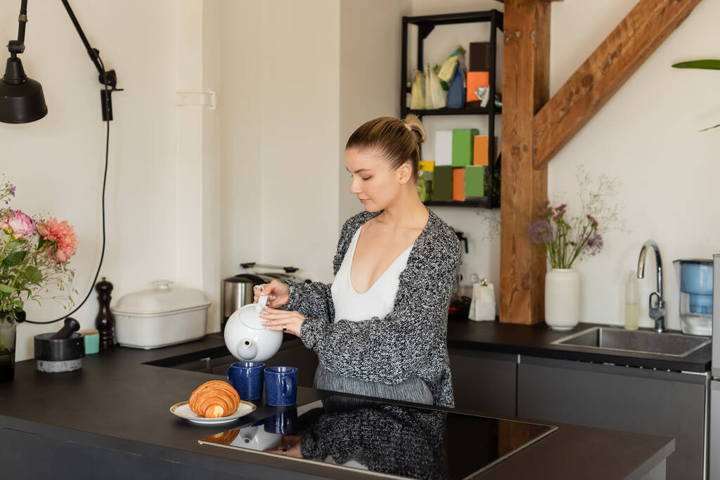 Junge Frau in Strickjacke gießt Tee in der Küche neben Croissant  - Foto, Bild