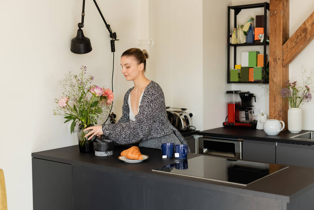 Woman in knitted cardigan putting vase with flowers near croissant and tea in kitchen  - Photo, Image