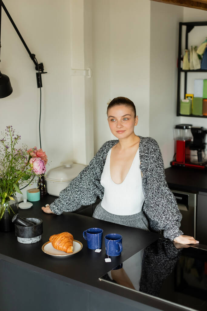 Mujer en punto cárdigan de pie cerca de croissant y tazas en la cocina  - Foto, imagen