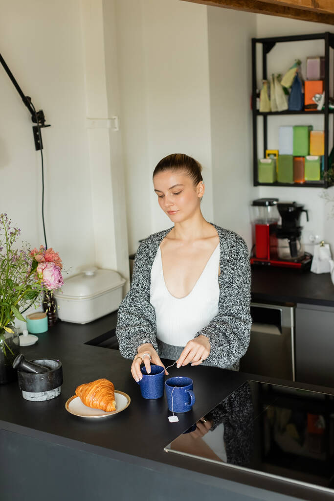 Frau in Strickjacke hält Tasse mit Teebeutel neben Croissant in Küche  - Foto, Bild