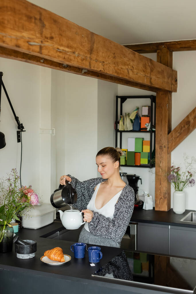 Uśmiechnięta kobieta wlewa wodę do czajnika w pobliżu smacznego rogalika i kubków w kuchni  - Zdjęcie, obraz