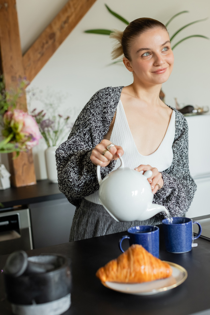 Positive Frau in Strickjacke gießt Tee in der Küche neben Croissant  - Foto, Bild