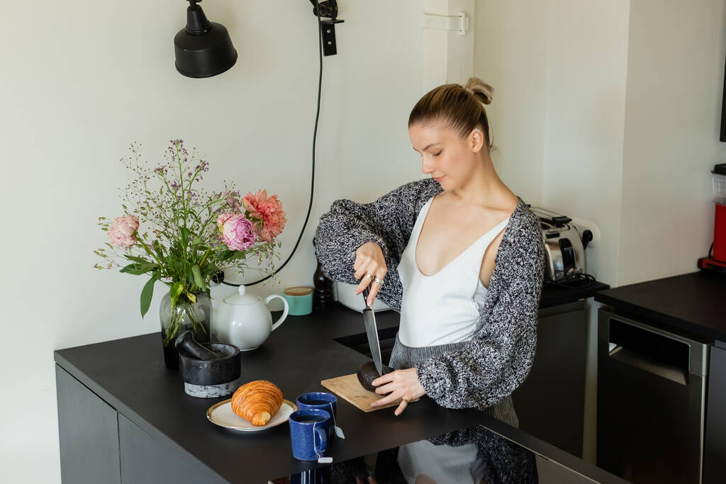 Frau in Strickjacke schneidet Avocado neben Croissant und Tassen in der Küche  - Foto, Bild