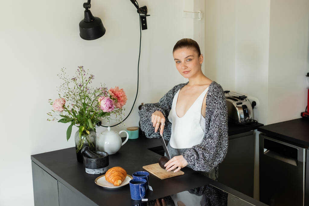 Jonge vrouw in warm vest snijden avocado in de buurt van thee en croissant in de keuken  - Foto, afbeelding