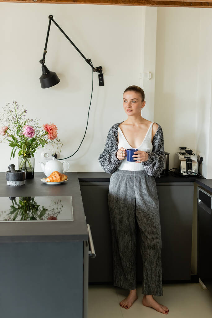 Θετική γυναίκα κρατώντας κύπελλο και κοιτάζοντας μακριά κοντά κρουασάν στη σύγχρονη κουζίνα  - Φωτογραφία, εικόνα