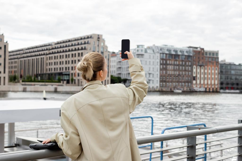 Back view of woman in trench coat taking photo on cellphone in Berlin  - Photo, Image