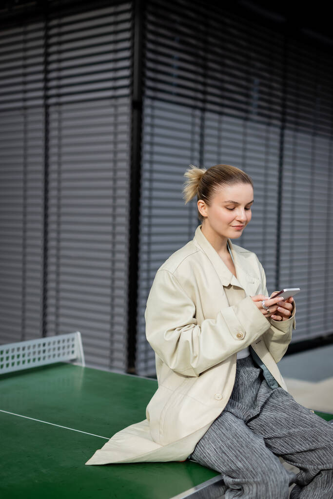 Junge Frau im Trenchcoat hält Smartphone in der Hand, während sie draußen am Tischtennistisch sitzt  - Foto, Bild