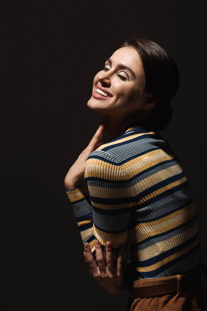 Χαρούμενη νεαρή γυναίκα με ριγέ ζιβάγκο χαμογελώντας απομονωμένη στο μαύρο  - Φωτογραφία, εικόνα
