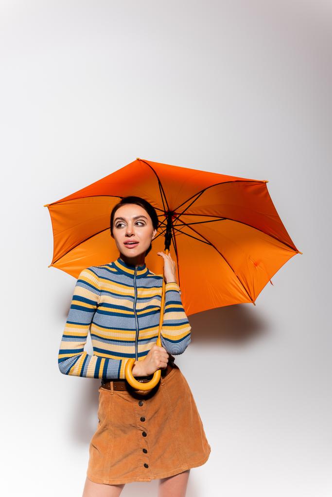 брюнетка в смугастій черепашці і спідниці, що стоїть під помаранчевою парасолькою на сірому
 - Фото, зображення
