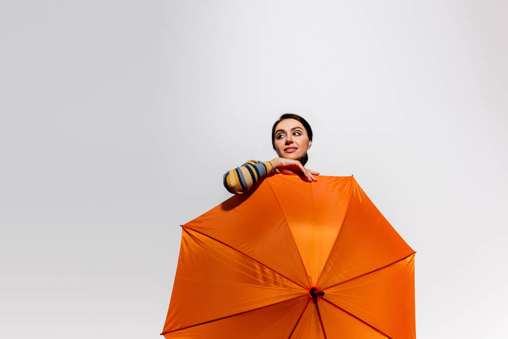 verträumte junge Frau posiert mit leuchtend orangefarbenem Regenschirm isoliert auf grau - Foto, Bild