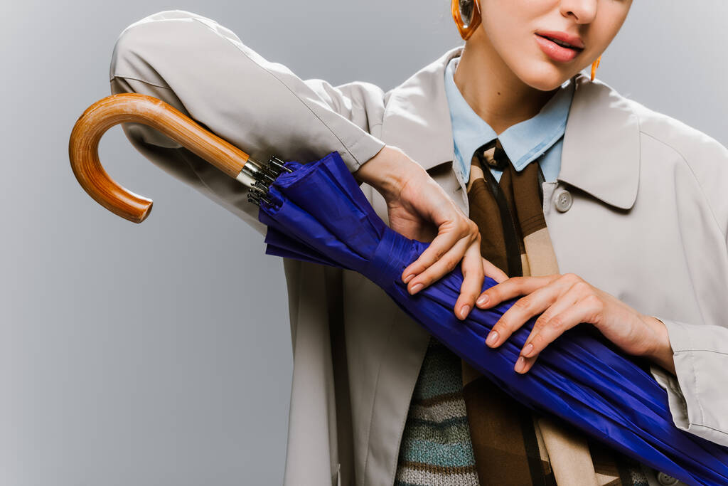 cropped view of young woman in trench coat posing with blue umbrella isolated on grey - Photo, Image