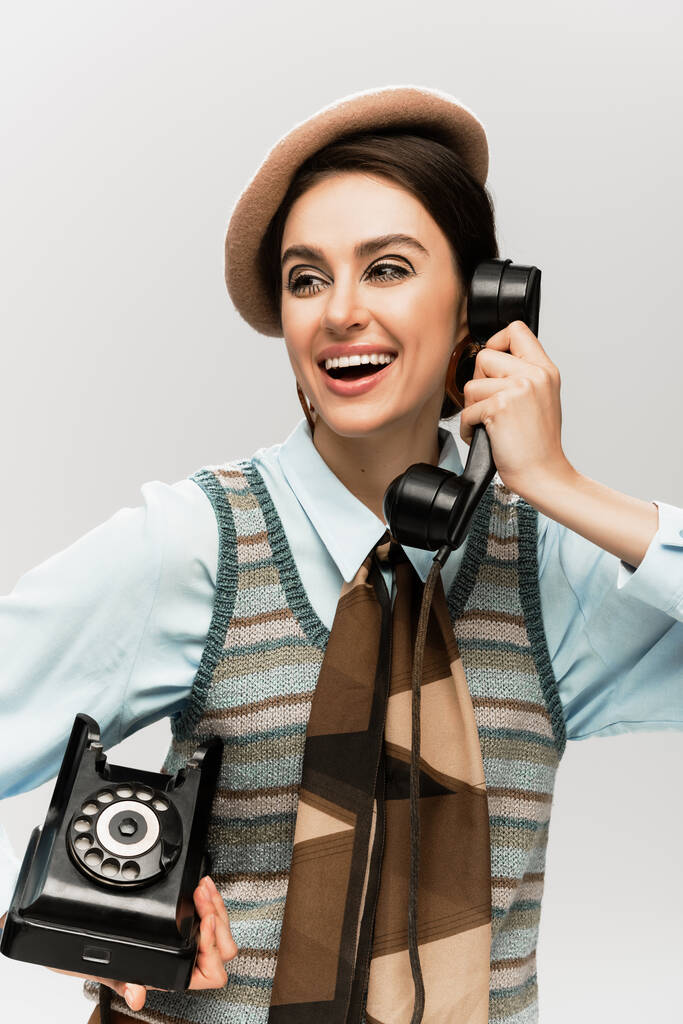 fröhliche junge Frau in Baskenmütze, die am alten Telefon vereinzelt auf grau spricht  - Foto, Bild