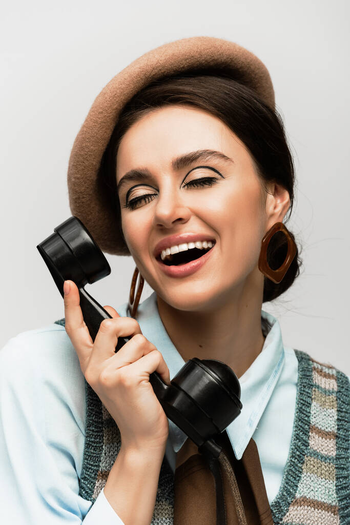 Lächelnde junge Frau mit Baskenmütze im Gespräch am Telefon, isoliert auf grau  - Foto, Bild