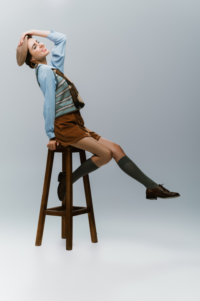 full length of joyful young woman in beret and autumnal clothes sitting on wooden high chair on grey - Photo, Image