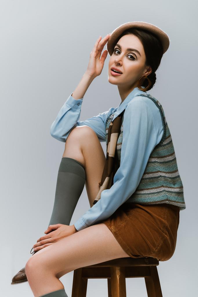 young woman in beret and autumnal clothes sitting on wooden high chair isolated on grey - Photo, Image
