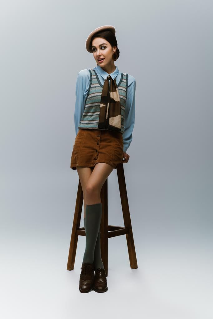 longitud completa de la mujer joven en boina y ropa otoñal de pie cerca de la trona de madera en gris - Foto, Imagen