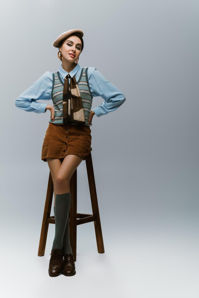 pełna długość młodej kobiety w berecie i jesiennej odzieży stojącej z rękami na biodrach w pobliżu drewnianego wysokiego krzesła na szarości - Zdjęcie, obraz