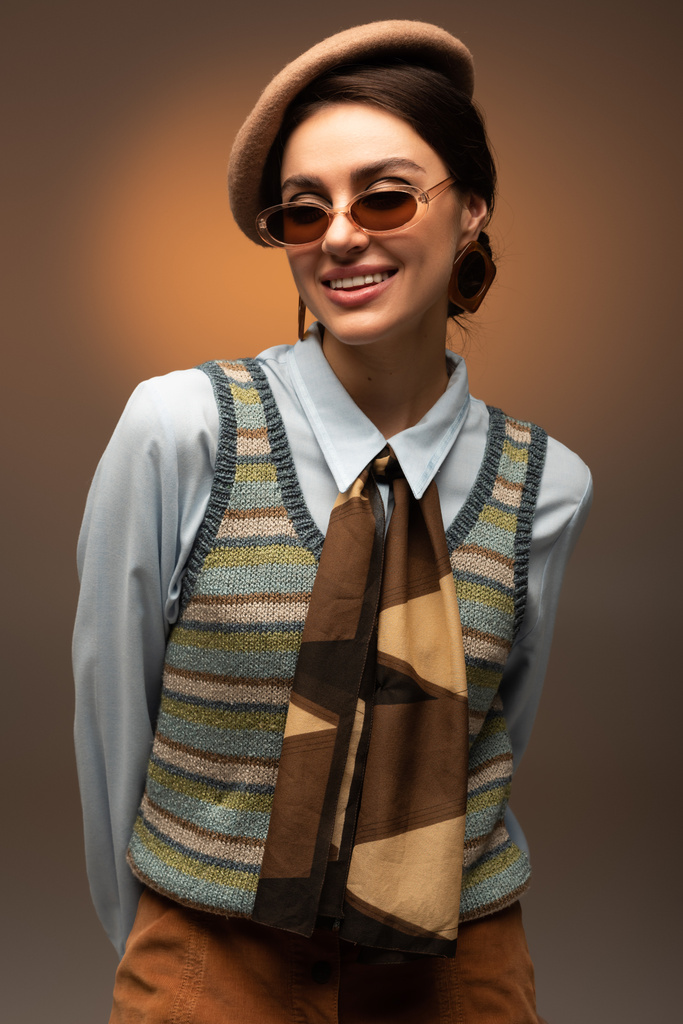 усміхнена молода жінка в береті і стильні сонцезахисні окуляри дивиться на камеру на коричневому
  - Фото, зображення