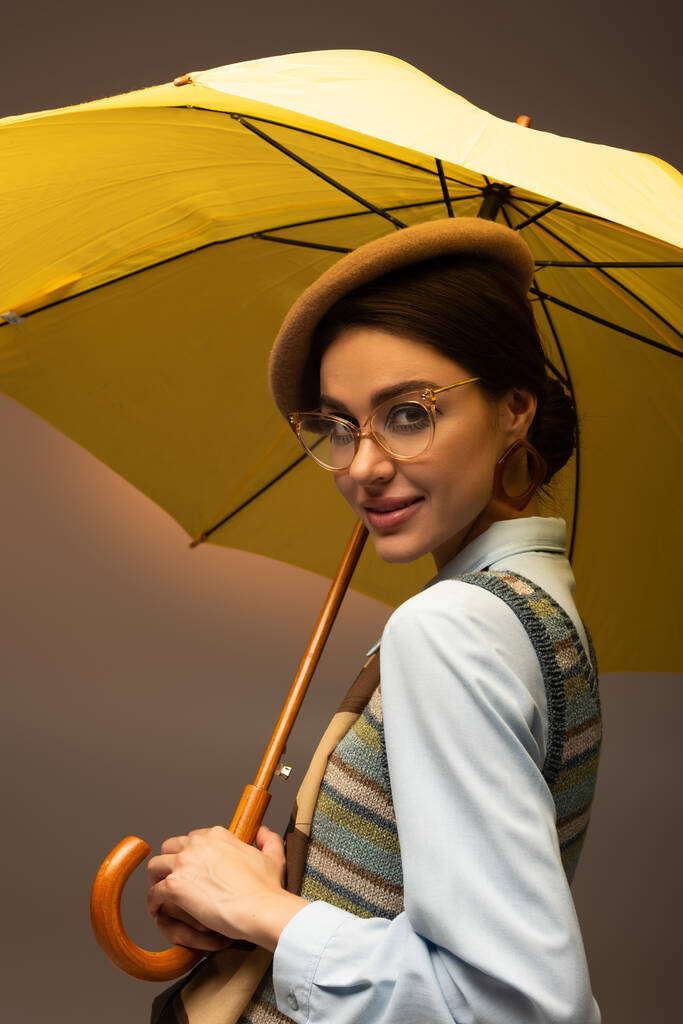 радісна молода жінка в береті і окулярах тримає жовту парасольку на сірому
 - Фото, зображення