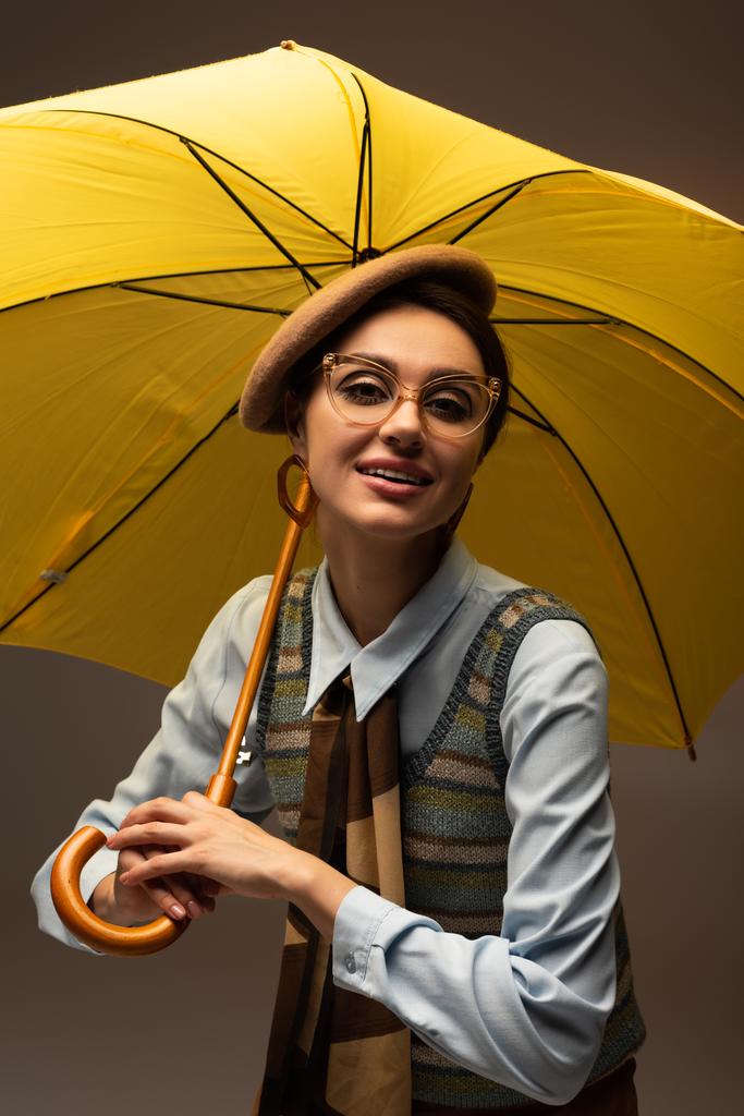 allegra giovane donna in berretto e occhiali con in mano ombrello giallo su grigio - Foto, immagini