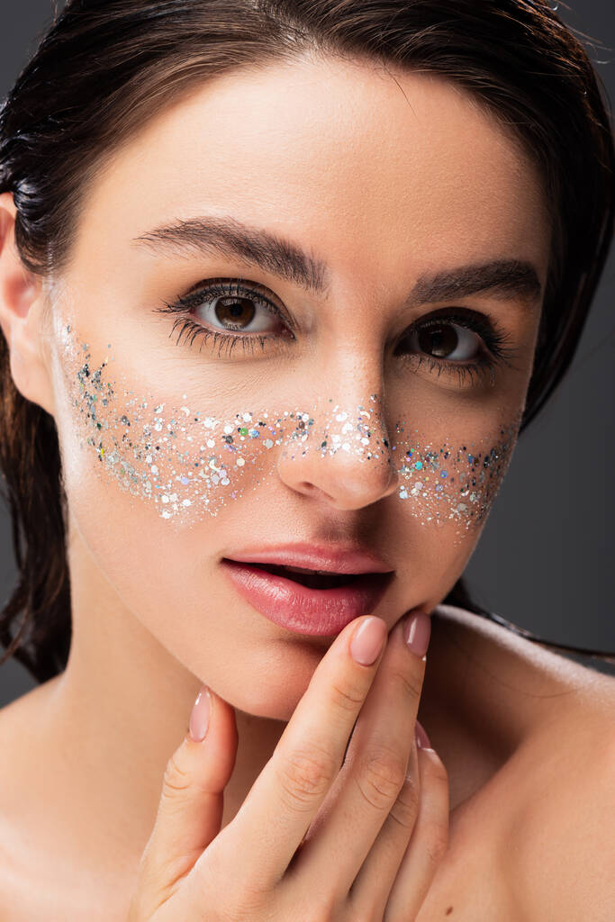 close up of young brunette woman with sparkling glitter on cheeks touching lips isolated on grey - Photo, Image