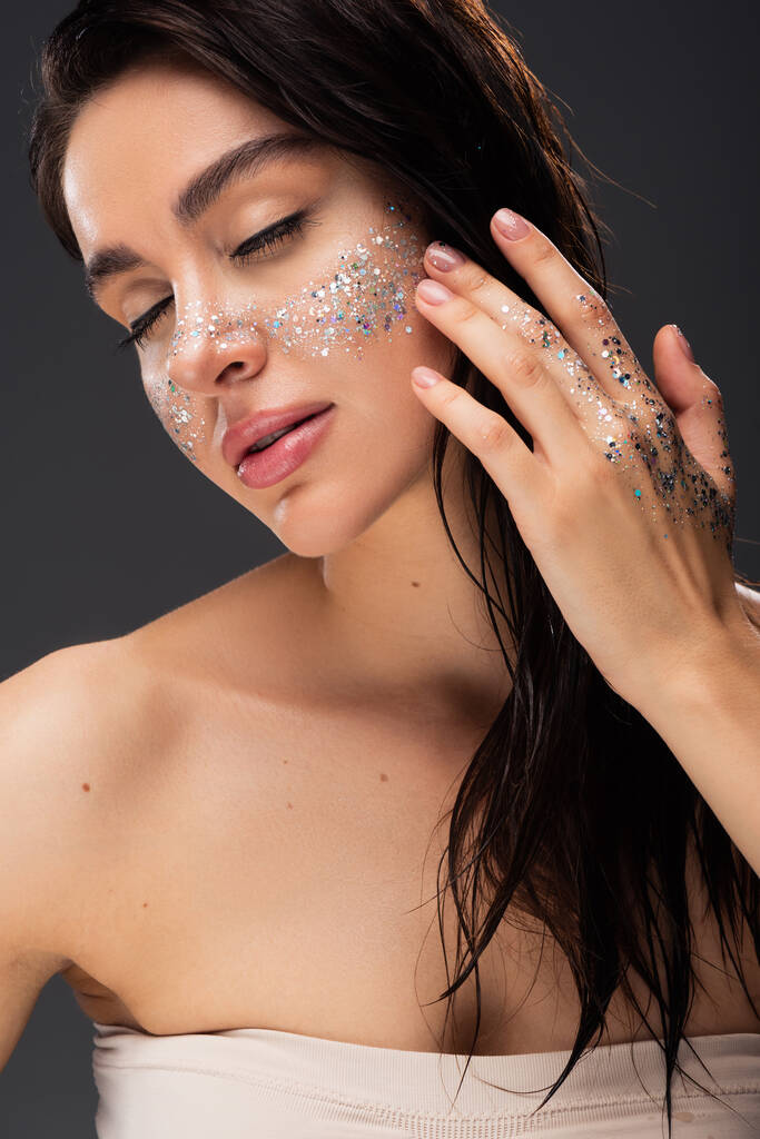 young brunette woman with closed eyes and sparkling glitter on cheeks and hand isolated on grey - Photo, Image