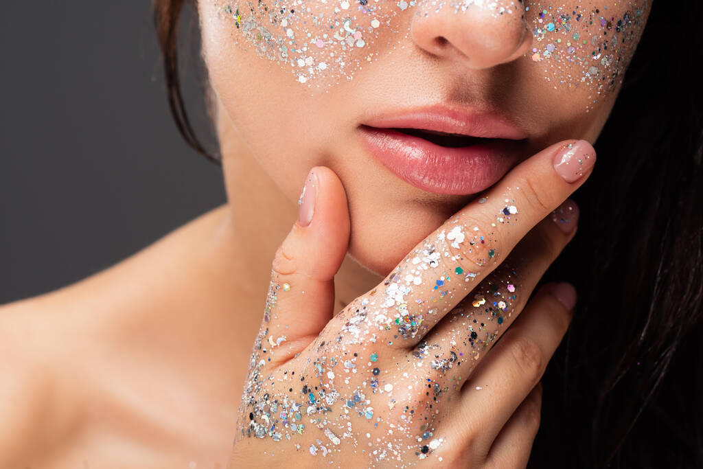 cropped view of young woman with sparkling glitter on cheeks and hand touching face isolated on grey - Photo, Image