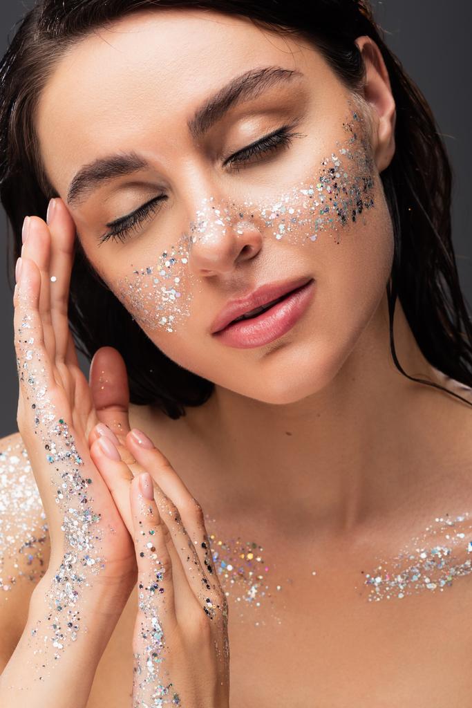portrait of sleepy young woman with sparkling glitter on cheeks and hands isolated on grey  - Photo, Image