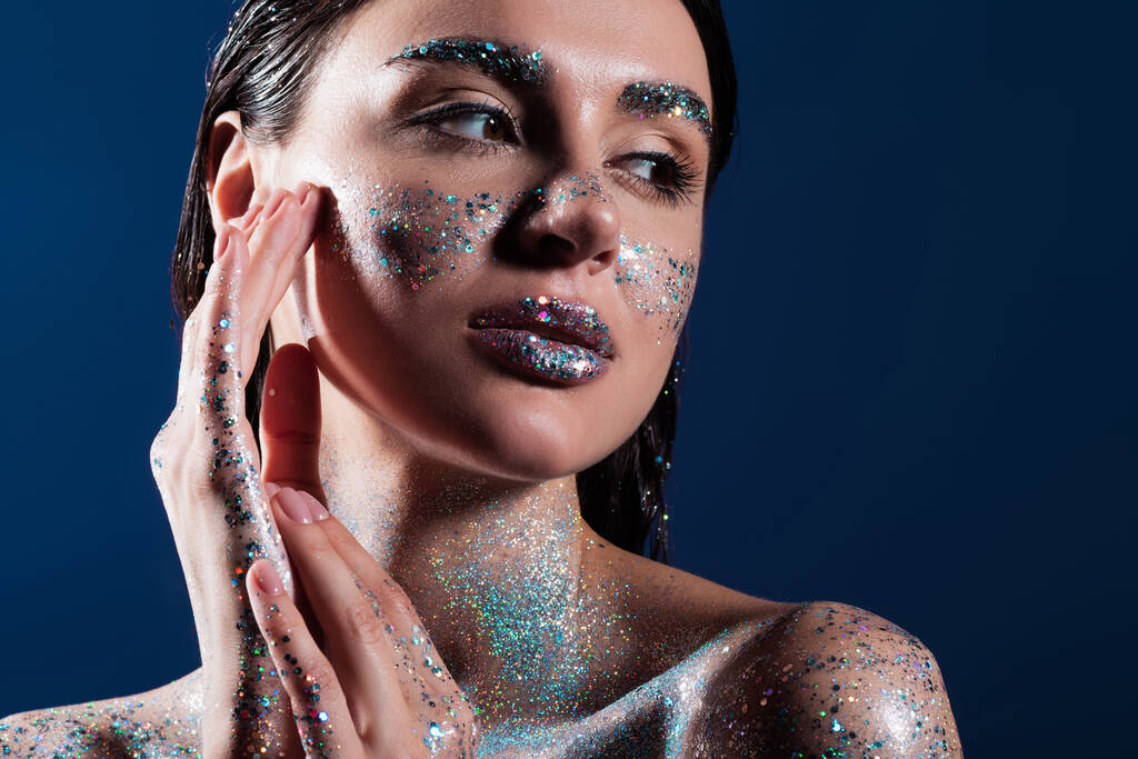 portrait of brunette young woman with glitter on body touching face isolated on blue  - Photo, Image
